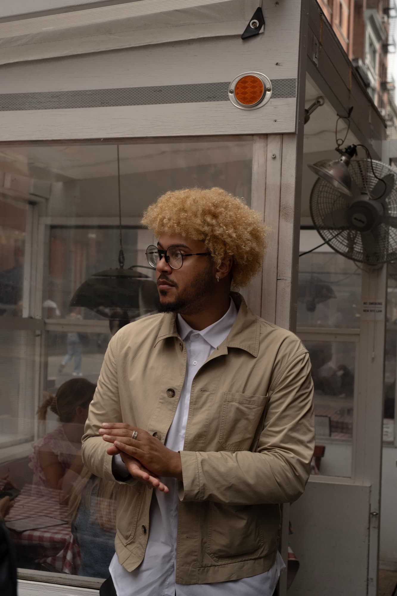 Ant with bleached curls and round glasses in a tan chore jacket outside a diner cart