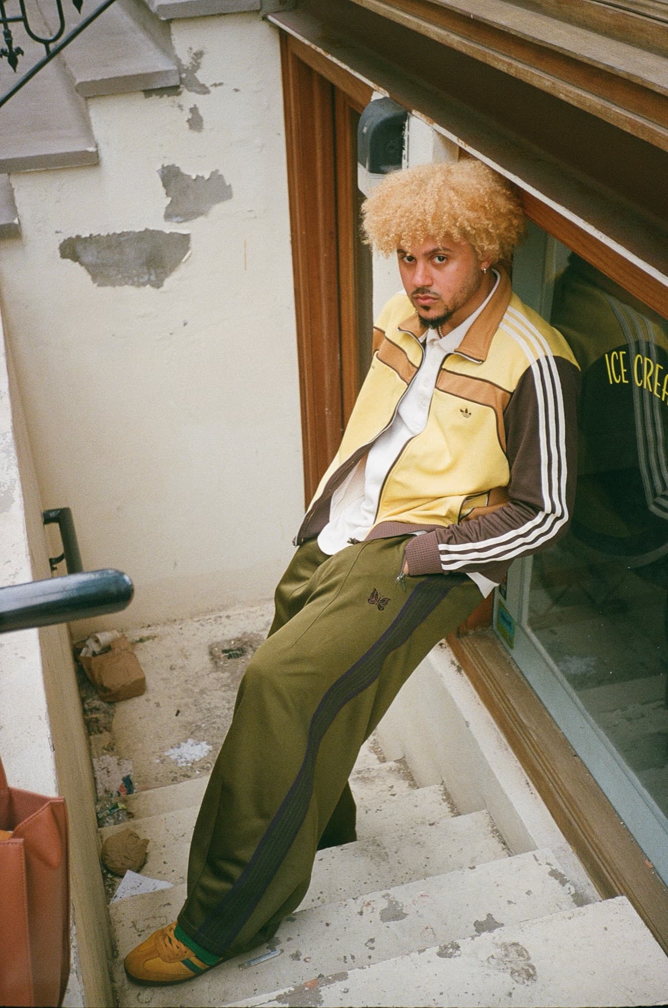 Ant leaning on a stairwell in a yellow and brown track jacket and olive track pants