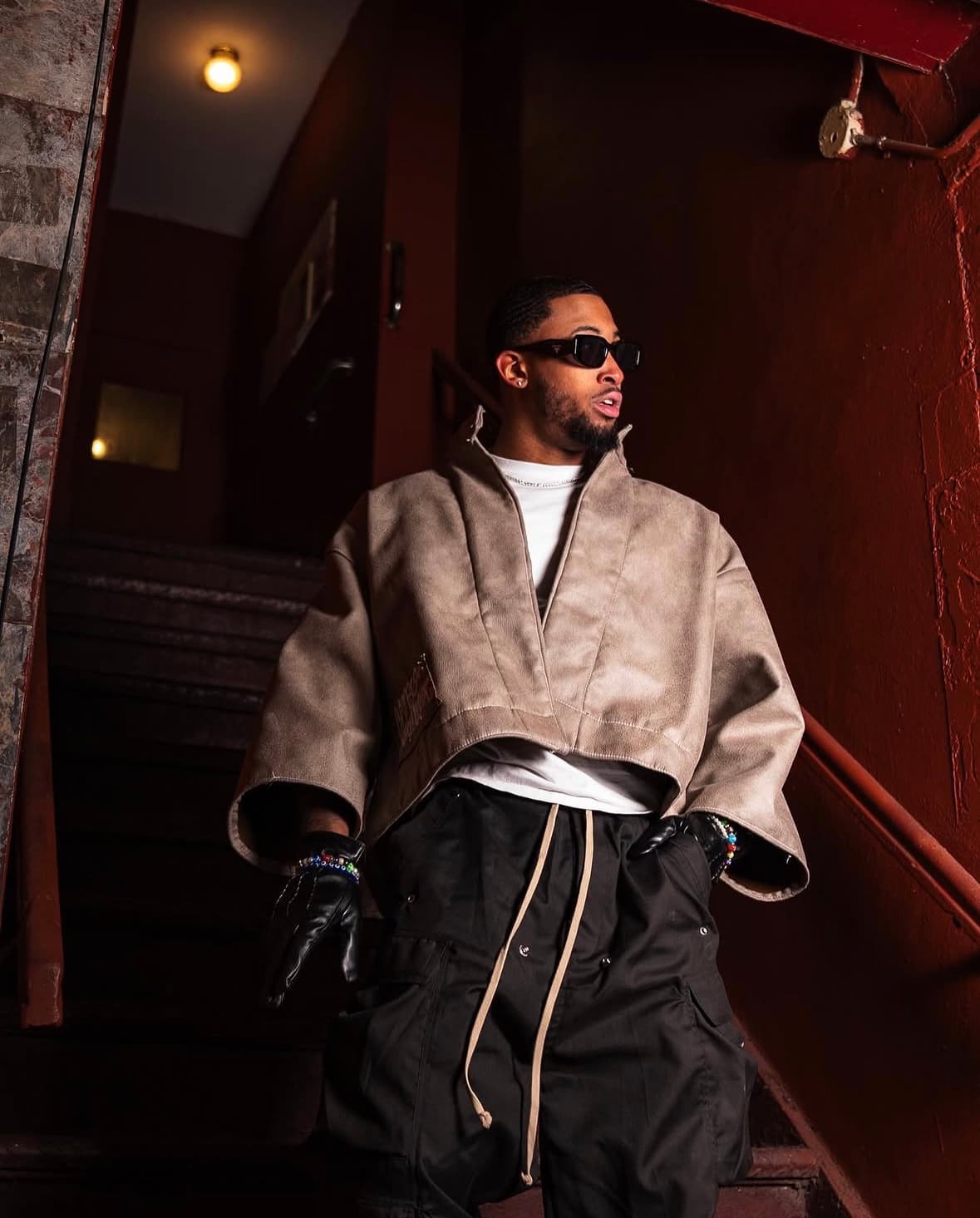 Bryce in a tan cropped jacket and sunglasses on a dim red stairwell