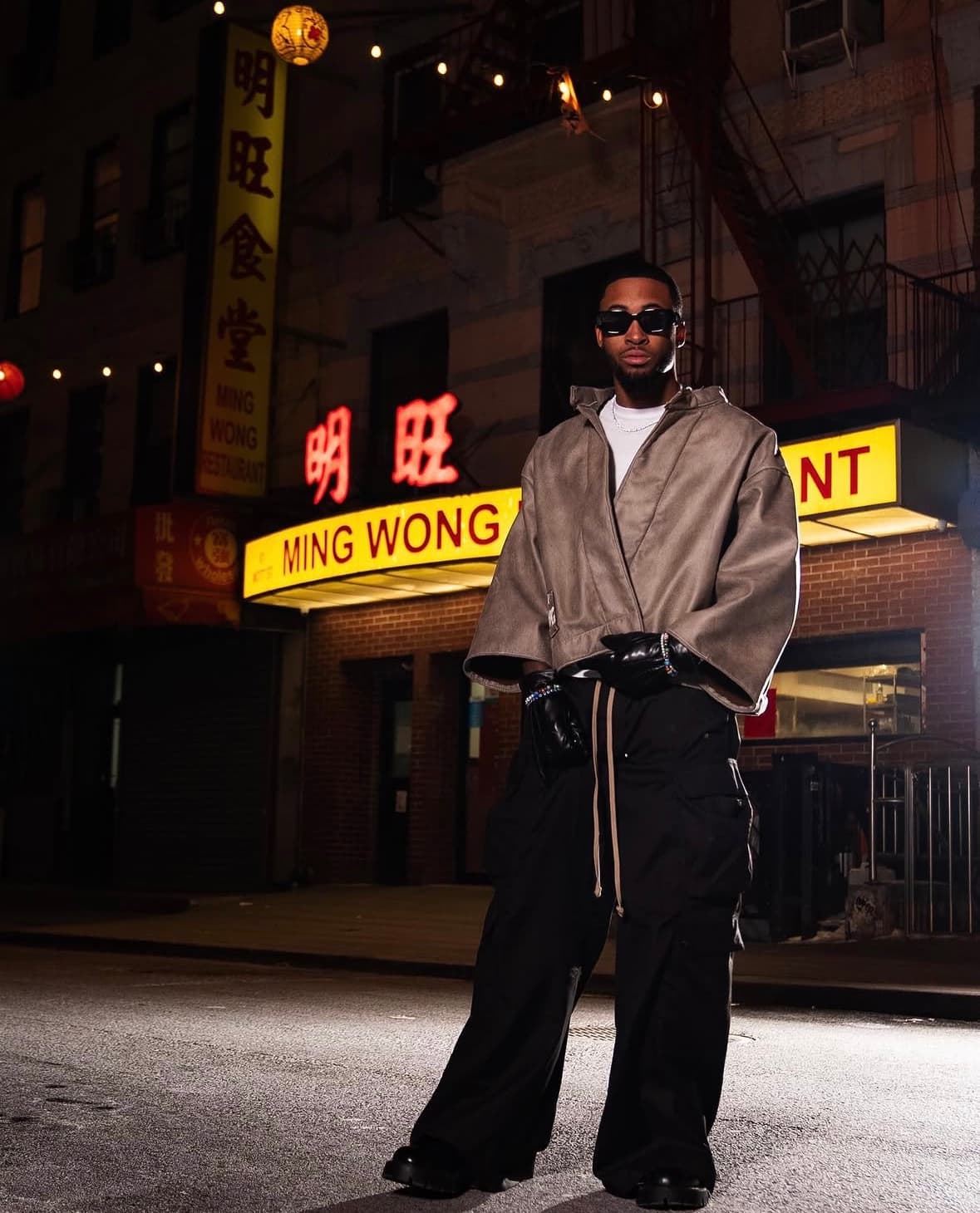 Bryce in a tan cropped jacket and wide black trousers under the Ming Wong neon sign at night