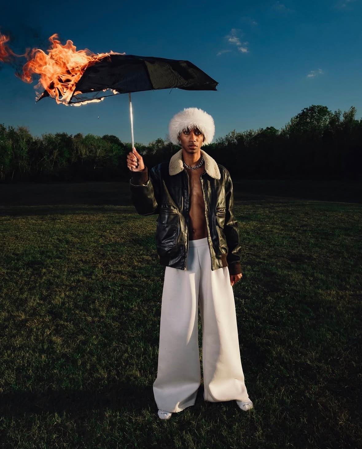 Dre in a white fur hat and leather jacket holding a burning umbrella in a field at dusk