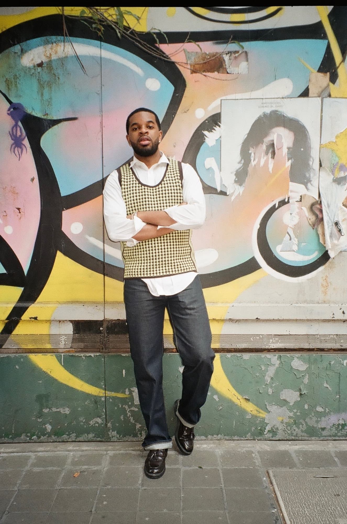 Khari in a knit vest and white shirt, arms crossed, in front of a graffiti wall