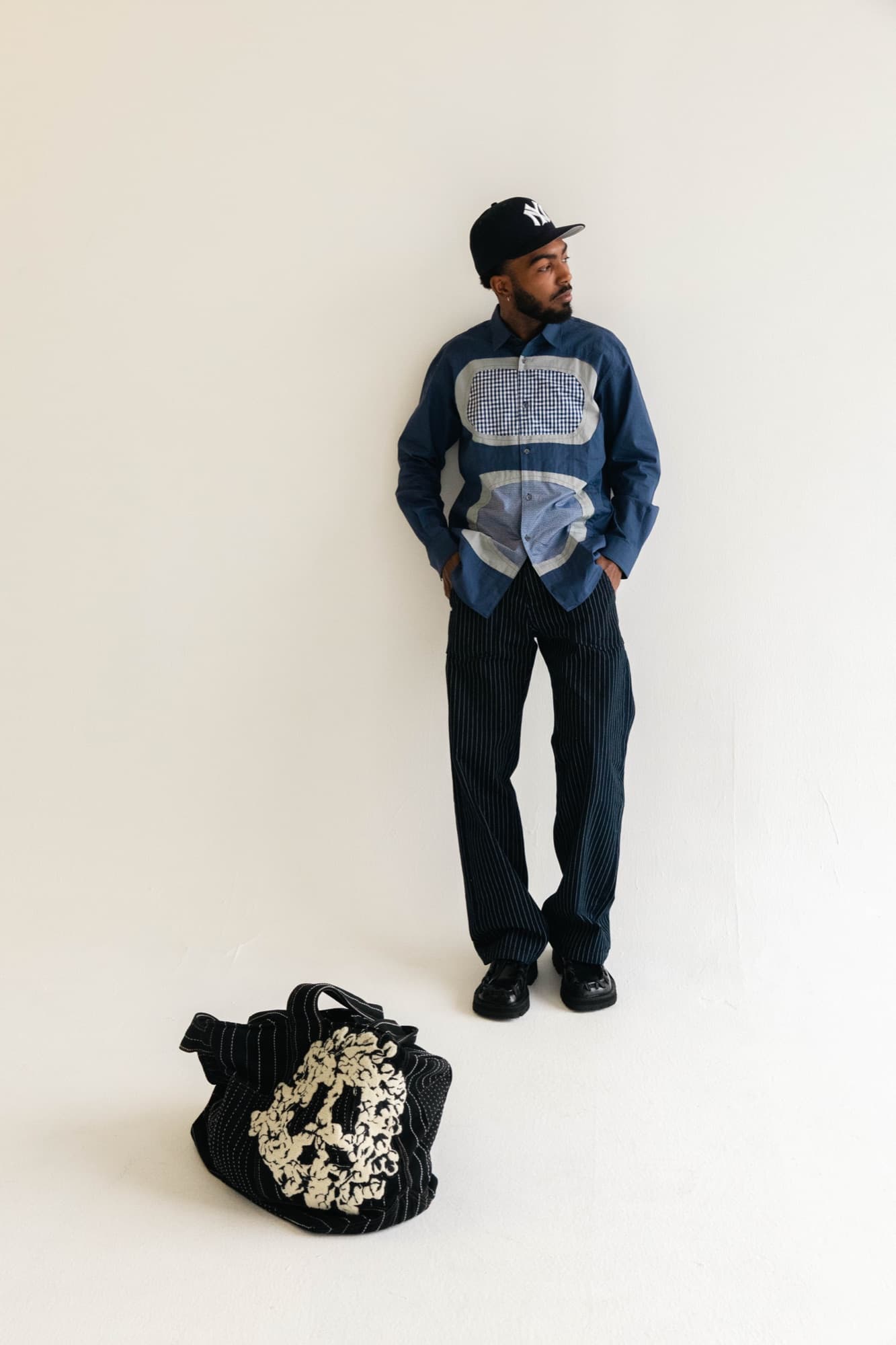 Khi in a patchwork blue shirt and pinstripe trousers, black cap, with an embroidered bag on the floor