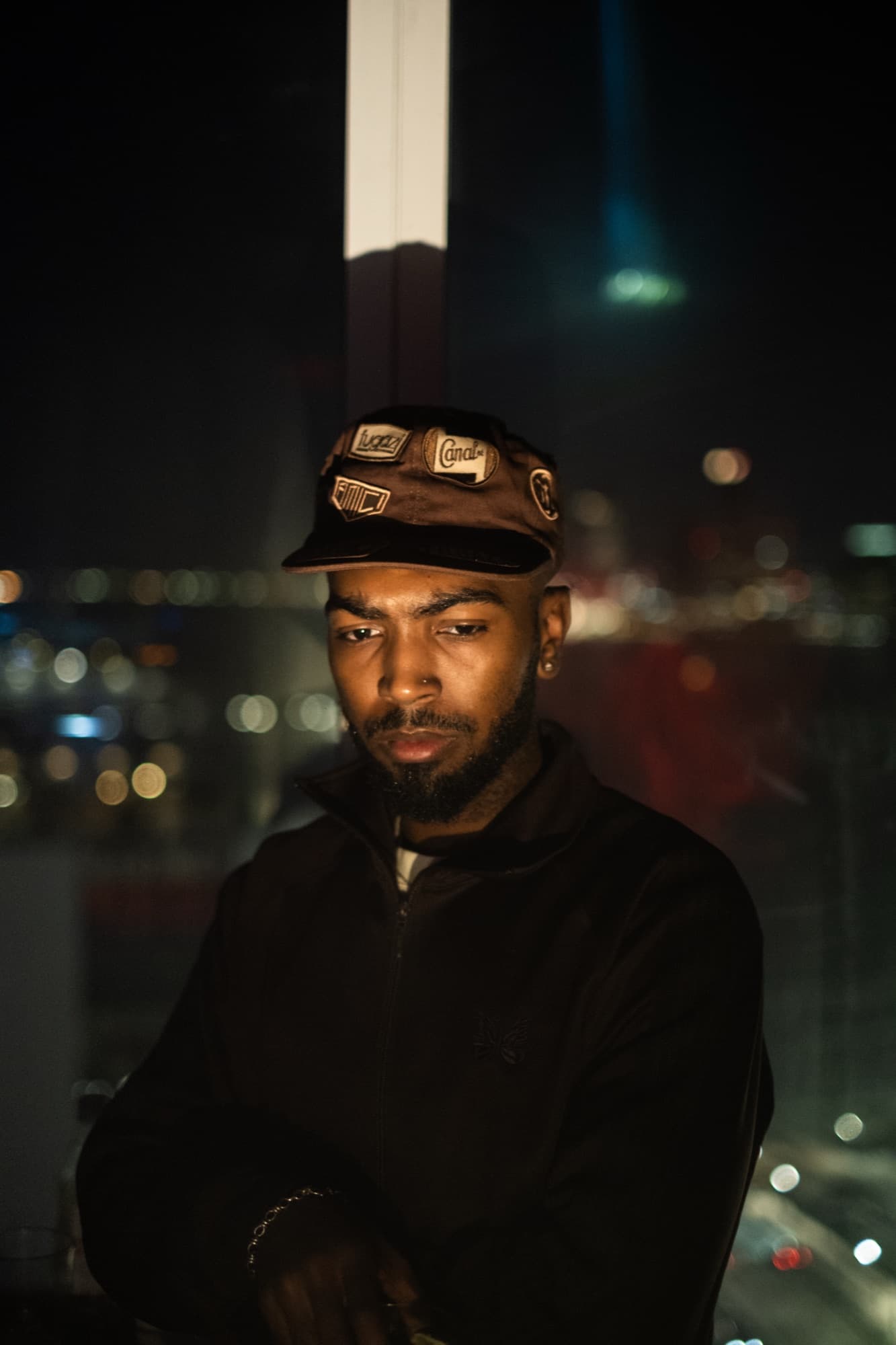 Khi at night in a patched brown cap and dark track jacket, city lights soft behind him