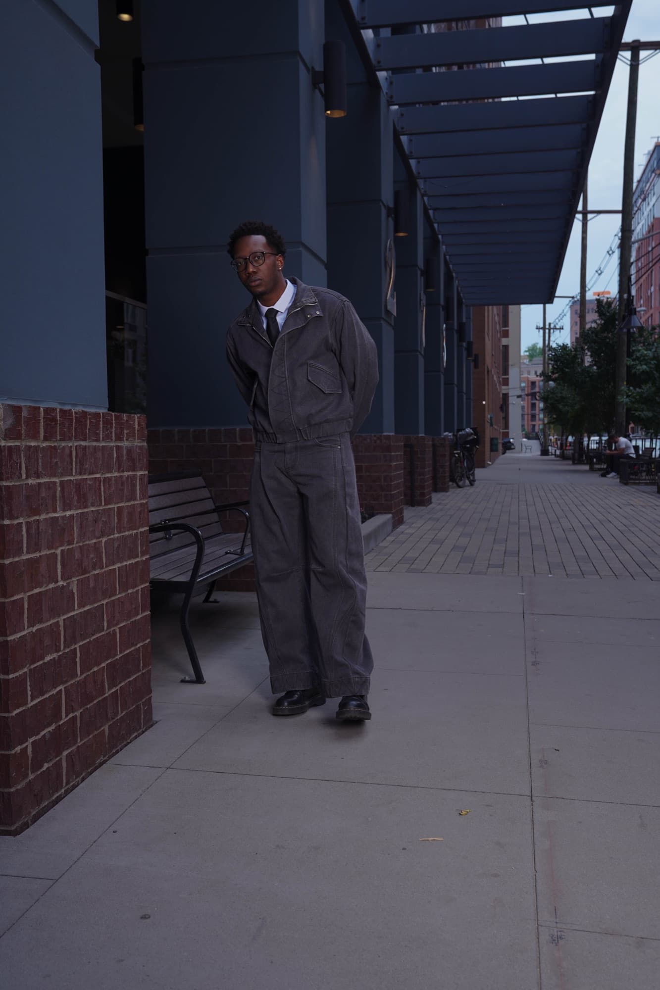 Stevo in a gray denim suit and tie walking along a brick colonnade at dusk